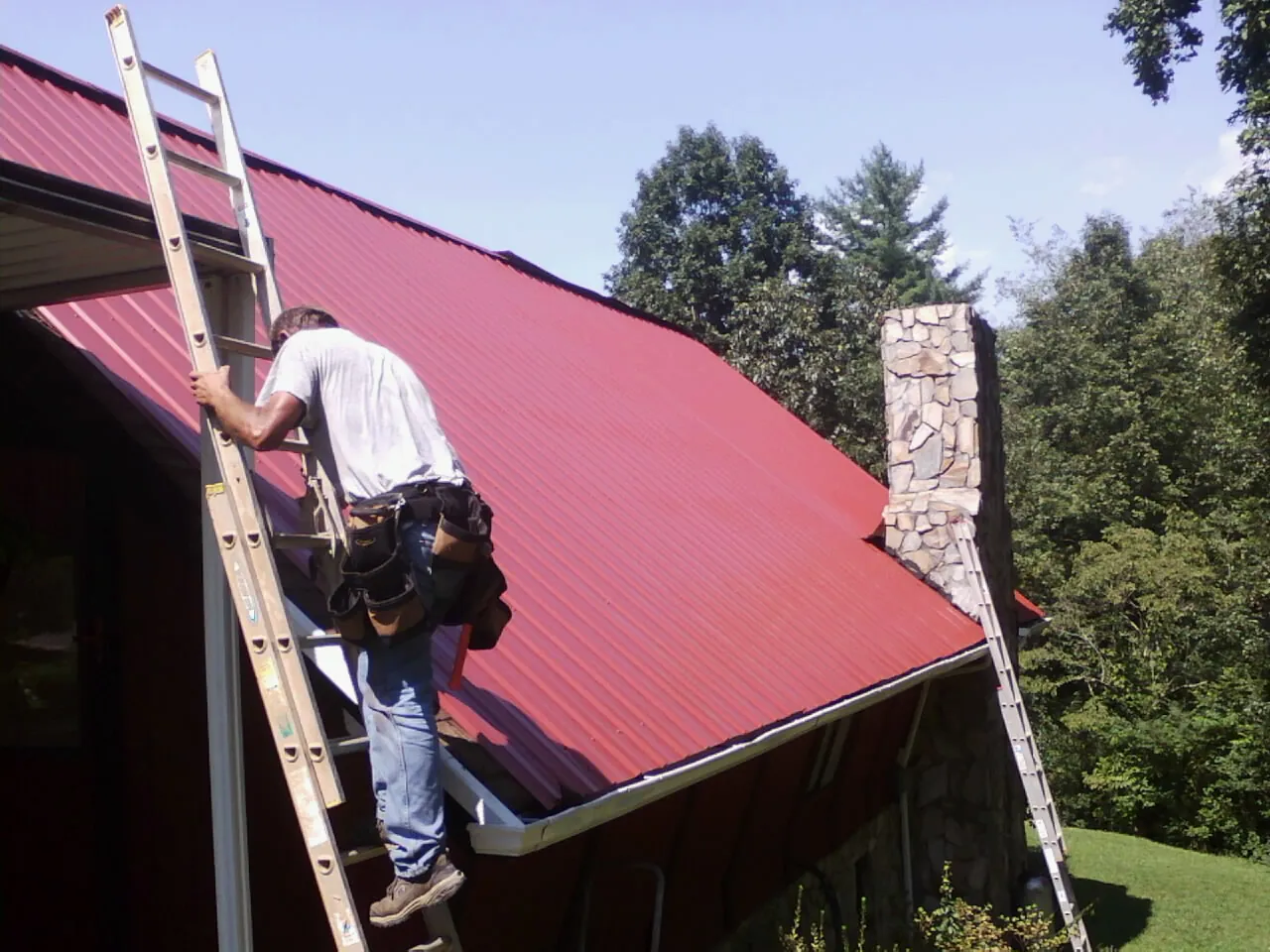 Roofer on a ladder beside a red metal roof and stone chimney — Soffit & Fascia Repair by Roofing Brothers in Jeannette